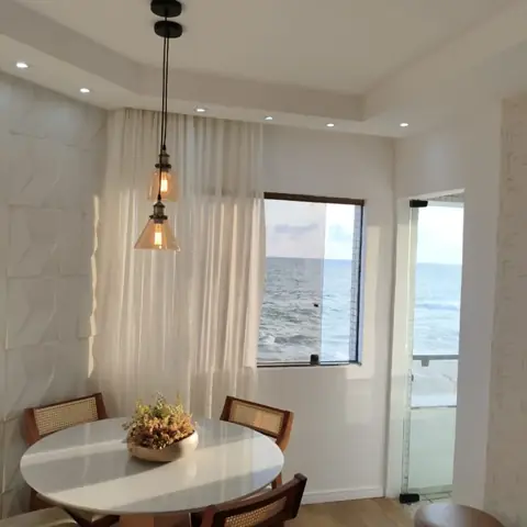 Mesa de jantar redonda ao lado da janela com vista para o mar, luminárias pendentes e parede com revestimento 3D